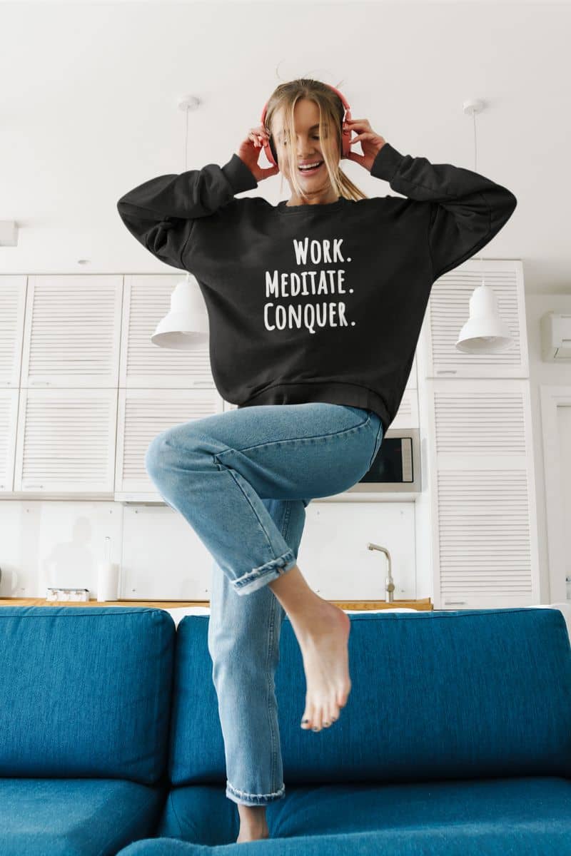 mindfulness sweatshirt with a person wearing a black sweatshirt with 'Work. Meditate. Conquer.' text, sitting on a blue couch in a kitchen.