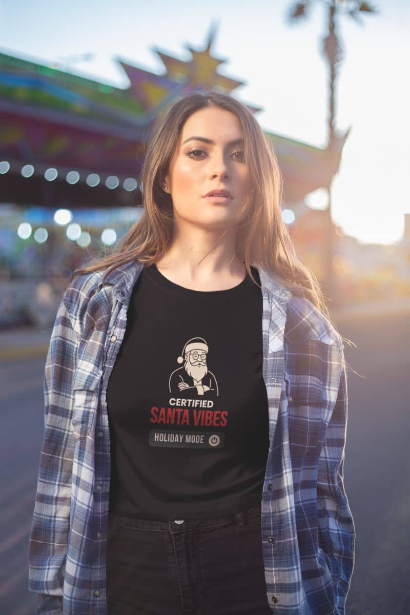 minimalist holiday shirt with a woman wearing a black t-shirt text 'Certified Santa Vibes – Holiday Mode On', standing in front of a colorful background.