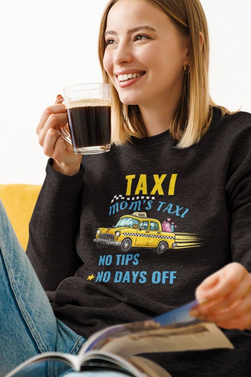 mom driver sweatshirt with a woman wearing a black sweater featuring the text 'Taxi, Mom’s Taxi – No tips, no days off', drinking coffee in the living room
