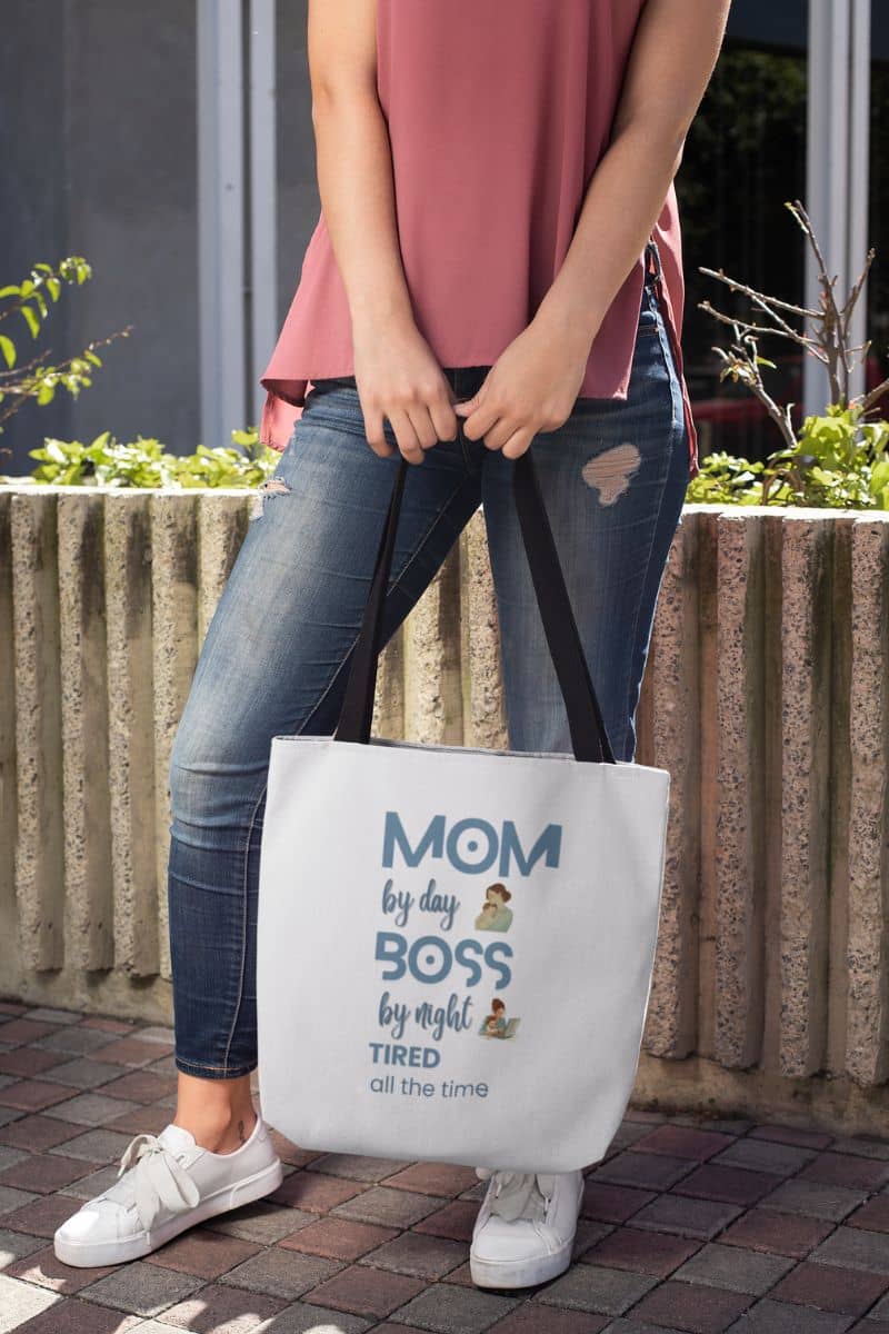 mom gift tote bag with a person holding a tote bag with text 'Mom by day, Boss by night, Tired all the time' outdoors.
