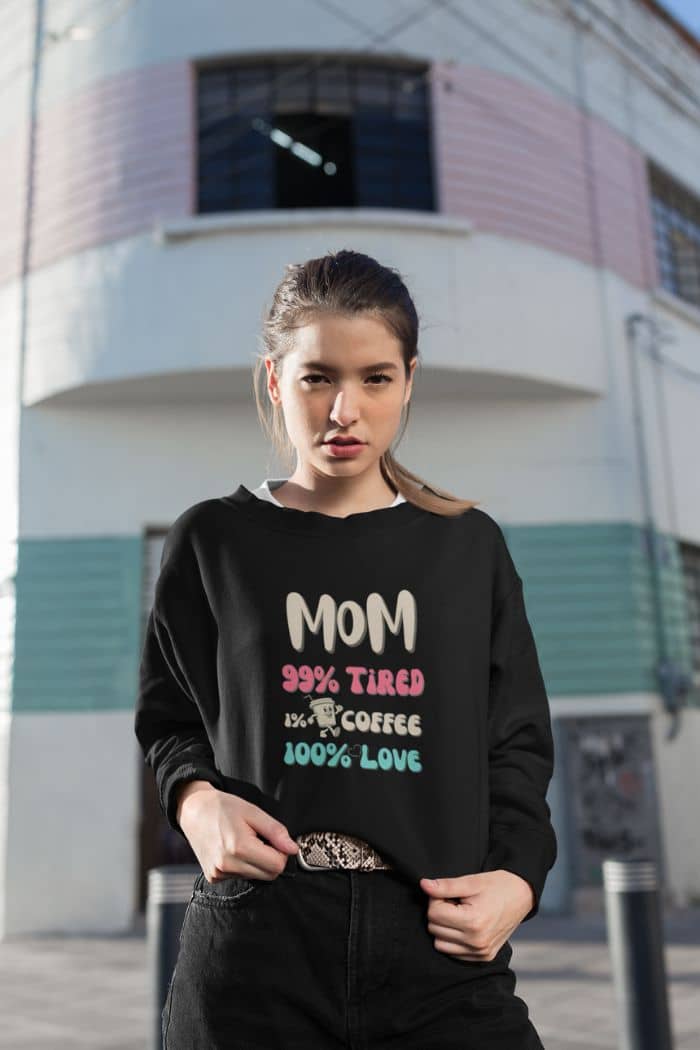 mom life sweatshirt with a woman wearing a black sweatshirt with text 'Mom 99% Tired, 1% Coffee, 100% Love' in front of a building