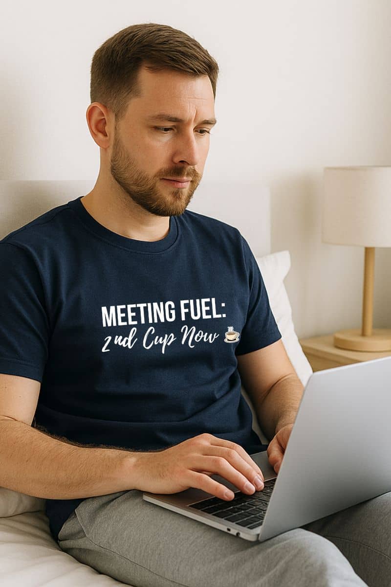 morning brew shirt with a man sitting on a bed using a laptop wearing a navy blue t-shirt with humorous text.