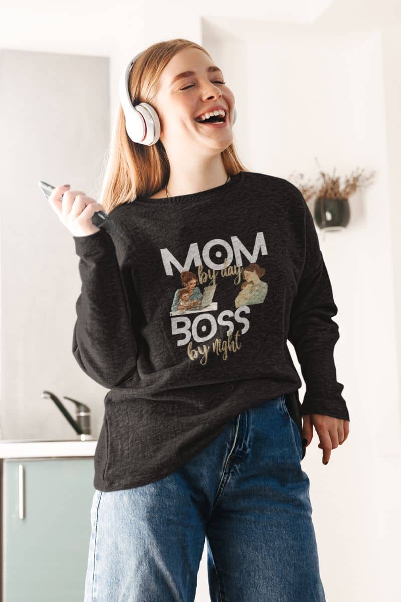 motherhood graphic sweatshirt with a woman wearing a sweatshirt featuring the text 'Mom by day, Boss by night' in a kitchen.