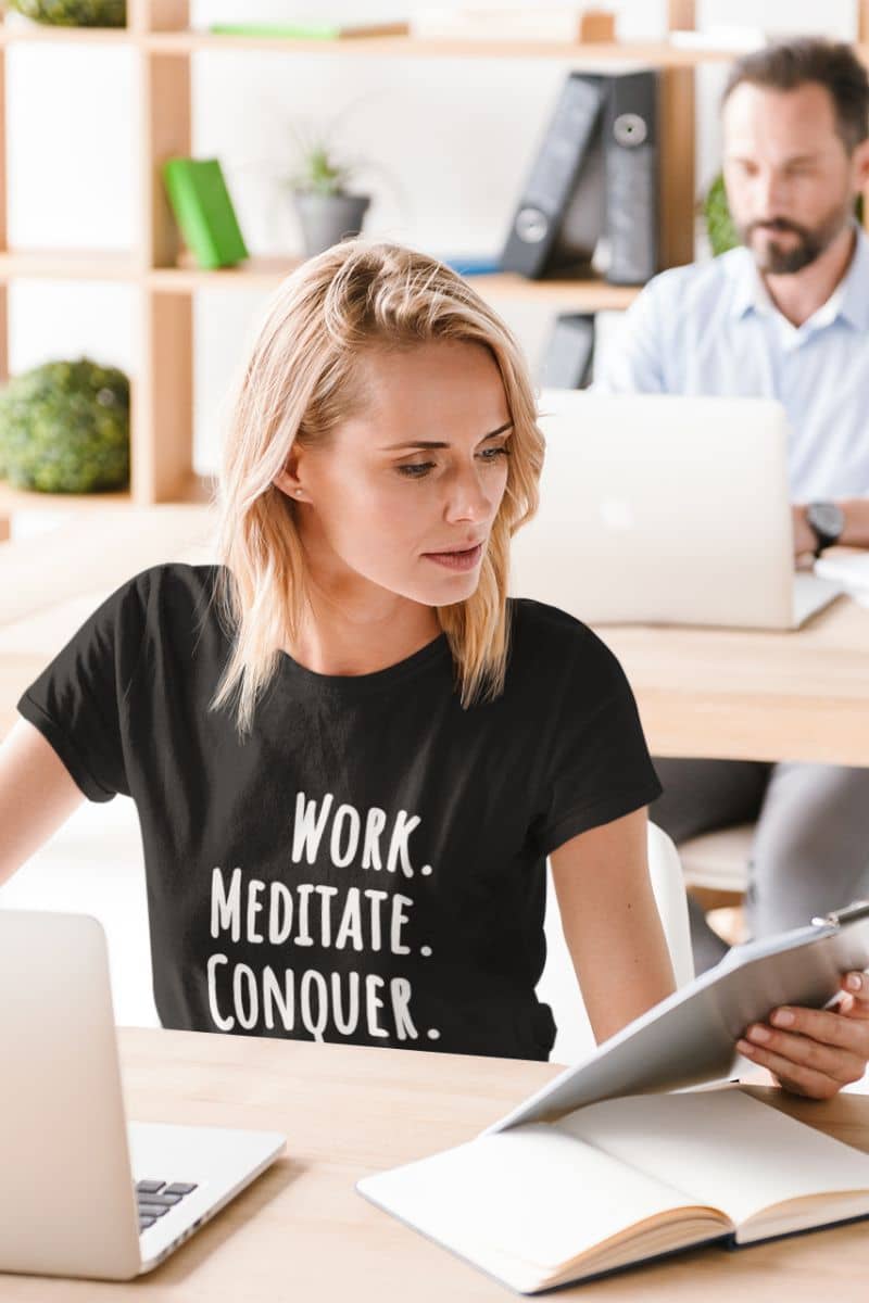 motivational graphic tee with a woman sitting at a desk with a laptop and tablet, wearing a black t-shirt with motivational text 'Work. Meditate. Conquer.'.
