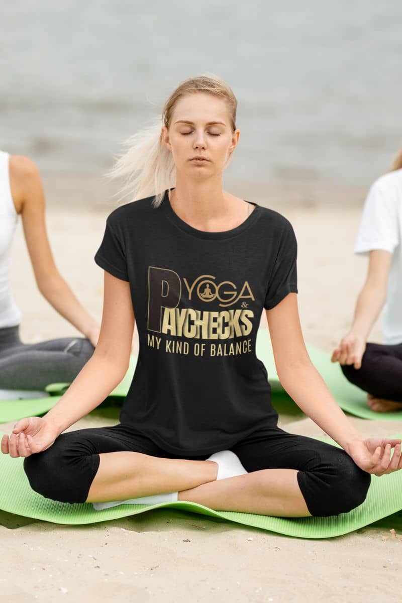 motivational work shirt with a woman sitting on a yoga mat with a black t-shirt featuring 'Yoga & Paychecks – My Kind of Balance' text.