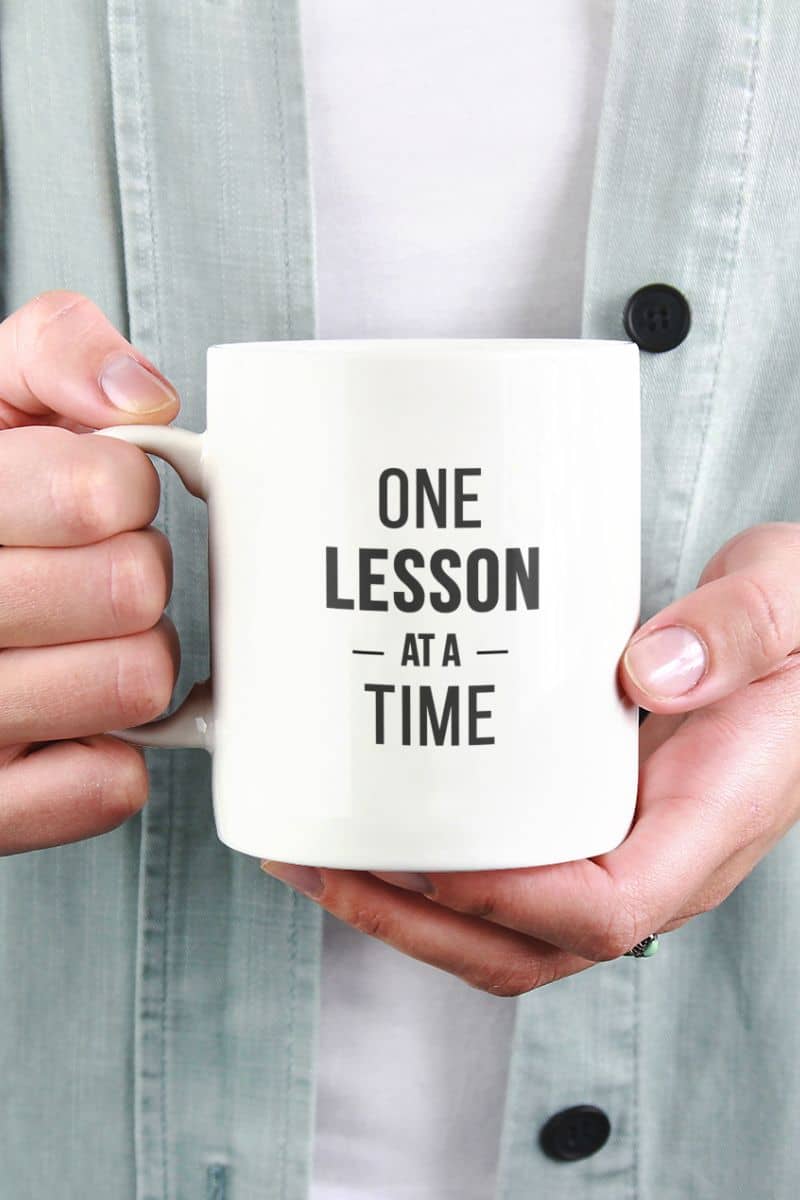 mug for teachers with a person holding a white ceramic mug with the text 'ONE LESSON AT A TIME' printed in black. The person is wearing a light blue button-up shirt over a white undershirt.
