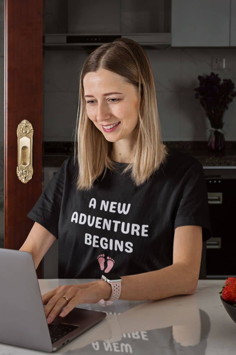 new parent graphic tee with a woman sitting at a table with a laptop, wearing a black t-shirt with text 'New Adventure Begins', in a kitchen setting.