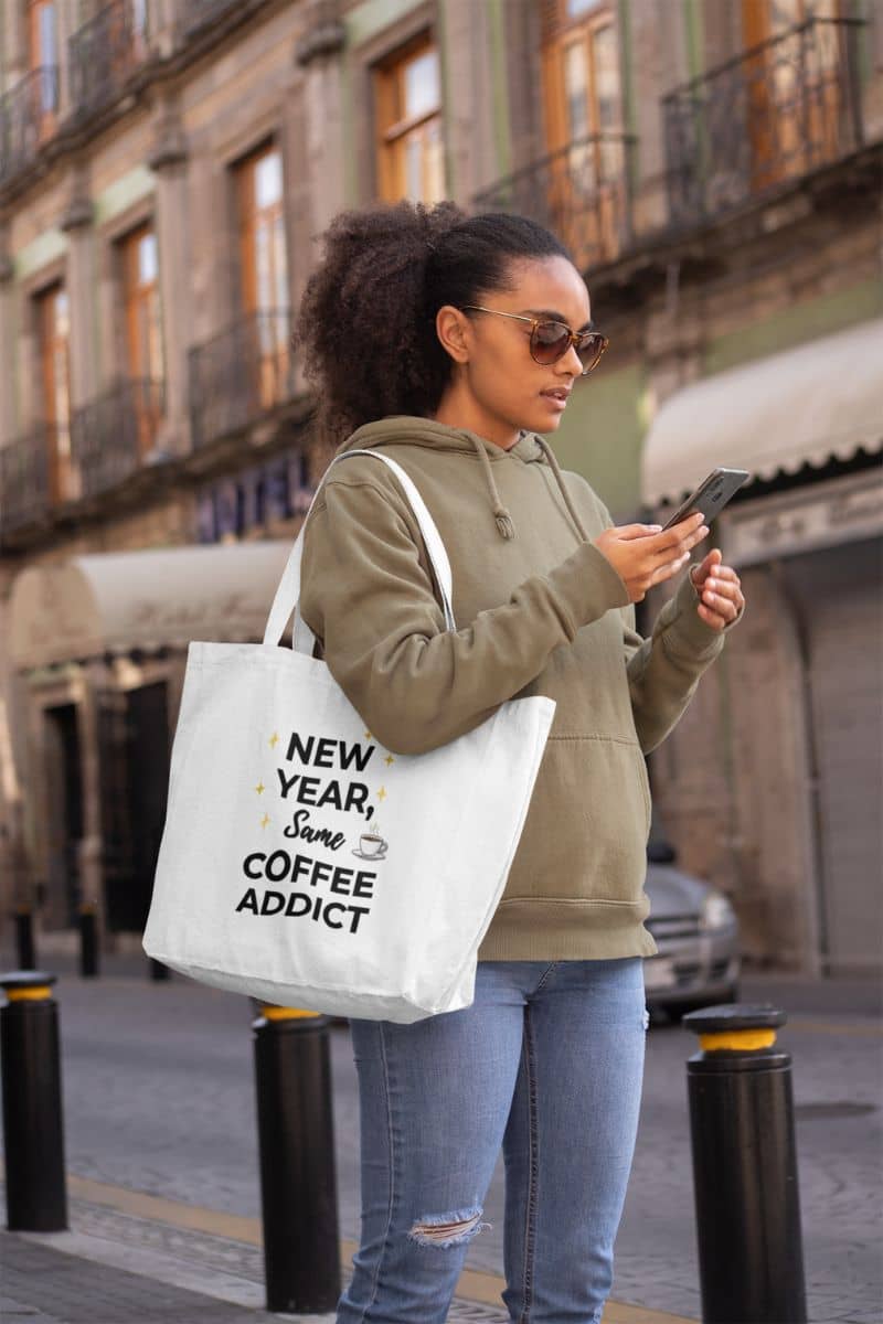 new year fashion bag with a woman carrying a tote bag featuring the text 'New Year, Same Coffee Addict', reading text messages on the street