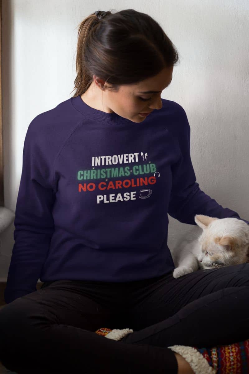 no caroling sweater with a person wearing a purple sweatshirt with text 'Introvert Christmas Club, No Caroling Please' sitting next to a white cat.