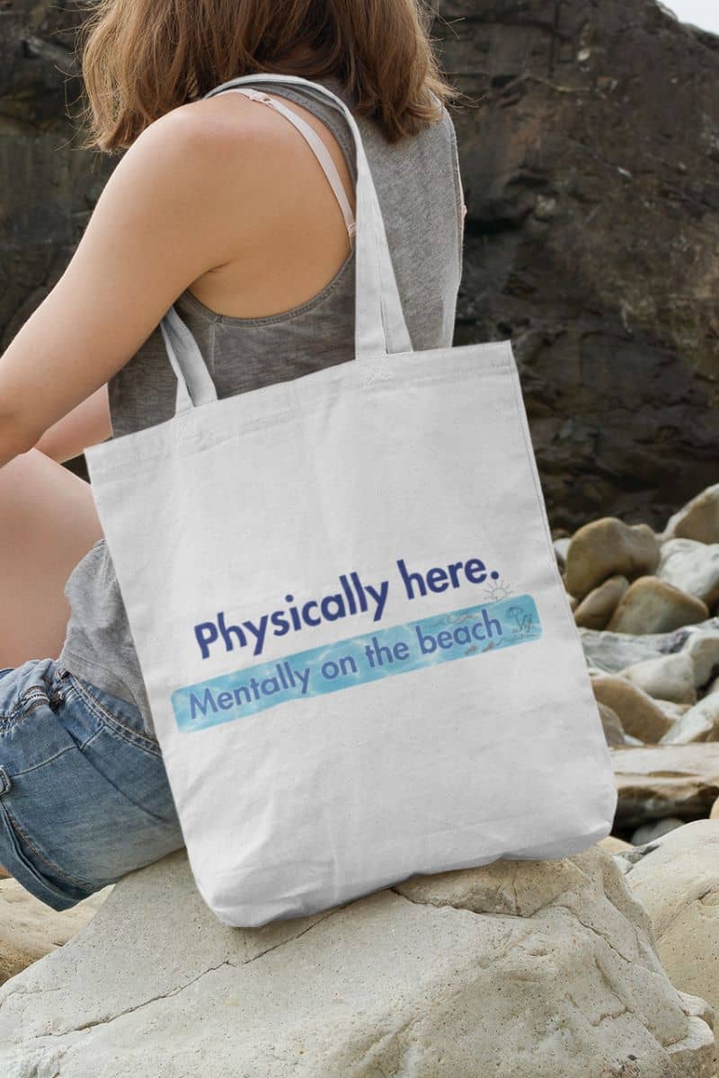 ocean vibes shopper bag with a person sitting on a rock by the beach with a tote bag that says 'Physically here. Mentally on the beach'.