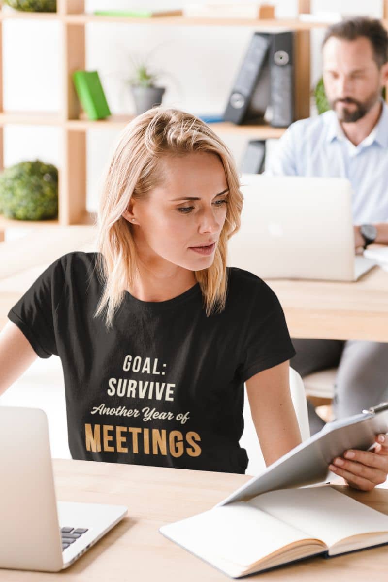 office burnout shirt with a woman in a black t-shirt with humorous text 'Goal: Survive Another Year of Meetings' sitting at a desk in an office setting.