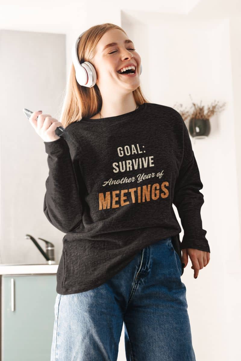 office burnout sweatshirt with a person wearing a sweatshirt with text 'Goal: Survive Another Year of Meetings', laughing in a kitchen.