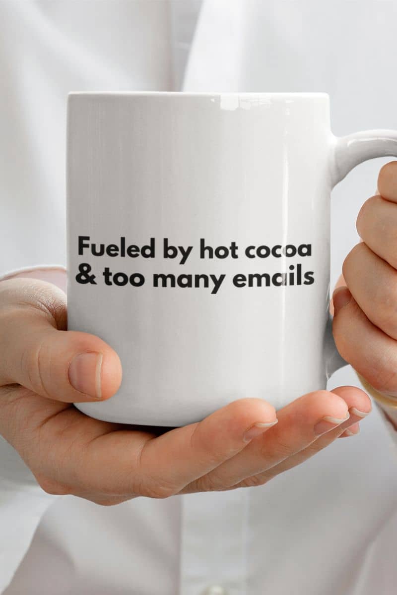 office humor coffee mug with a white mug held by hands with text 'Fueled by hot cocoa & too many emails' on a white background