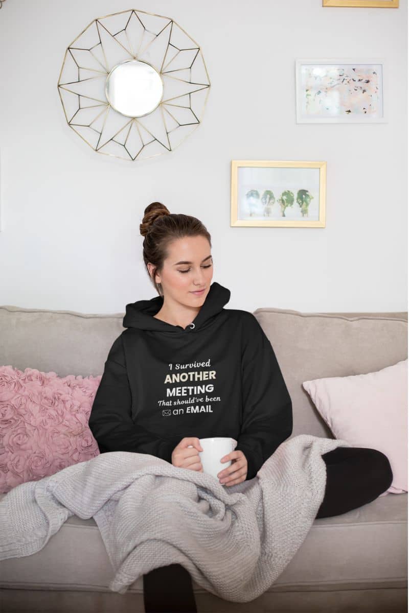 office humor hoodie with a woman sitting on a couch wearing a black hoodie with text 'I Survived Another Meeting That Should’ve Been an Email', holding a mug.