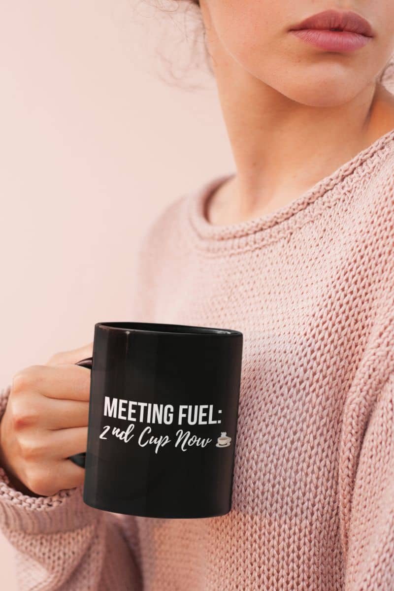 office humor mug with a person holding a black mug with text 'Meeting Fuel: 2nd Cup Now', wearing a pink sweater.