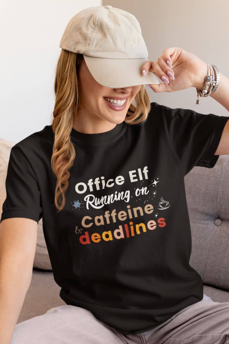 office humor shirt with a woman wearing a black t-shirt with text 'Office Elf, Running on Caffeine & Deadlines', sitting on a couch.