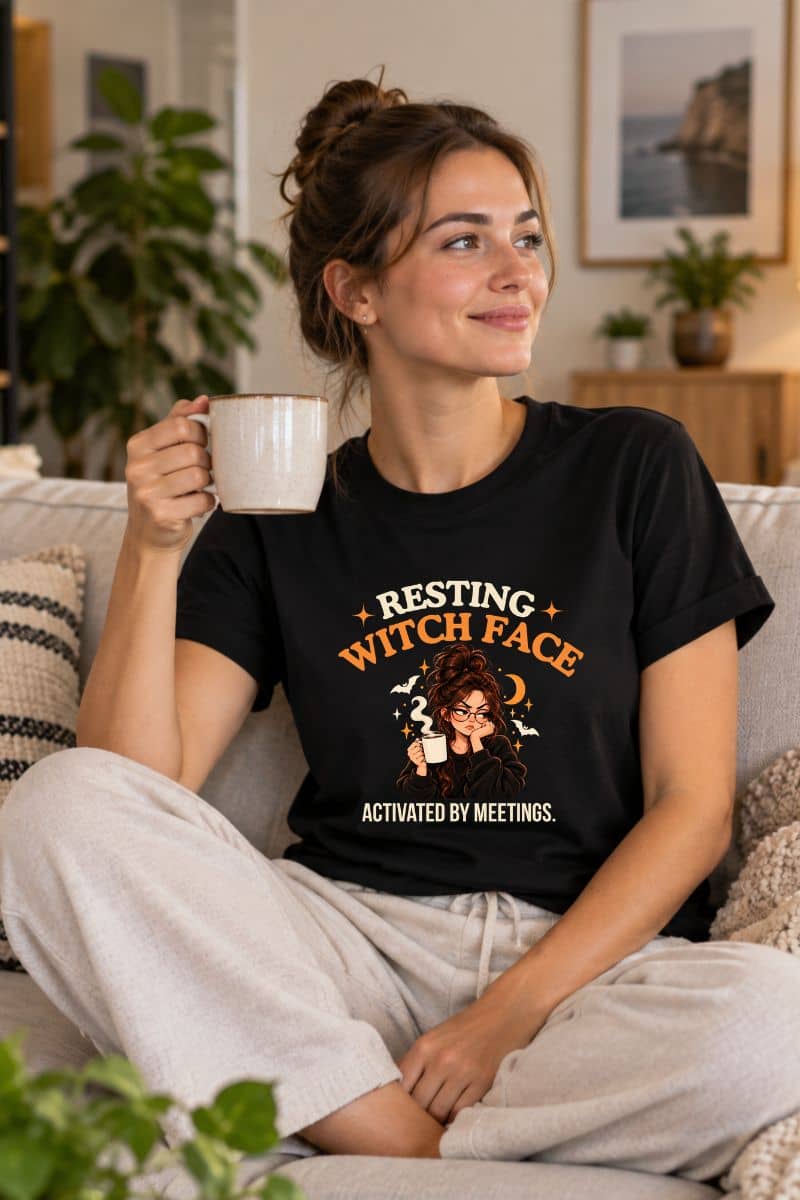 A woman drinking coffee on a sofa, wearing an office humor shirt featuring a “Resting Witch Face. Activated by Meetings.” graphic with a woman’s face.