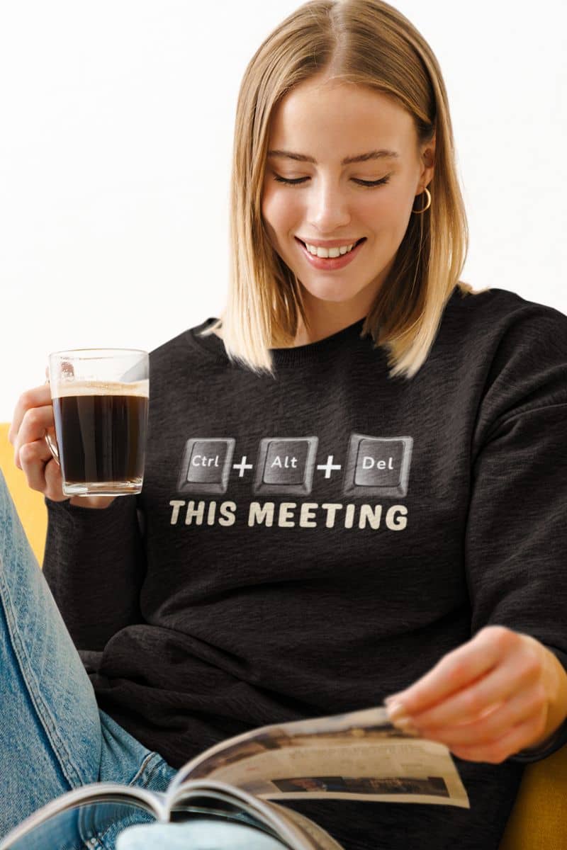 office humor sweatshirt with a woman wearing a black sweatshirt with a humorous computer keyboard design and text 'Ctrl + Alt + Del This Meeting', holding a cup of coffee and reading a newspaper.