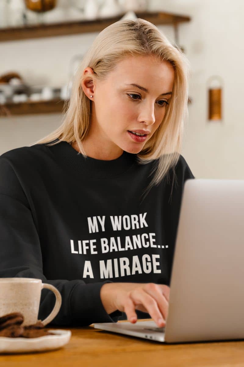 office humor sweatshirt with a woman in a black sweatshirt with text 'My Work-Life Balance Is a Mirage' working on a laptop in a kitchen.