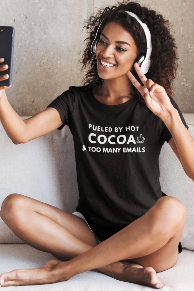 office humor t-shirt with a woman wearing a black t-shirt with text 'Fueled by hot cocoa & too many emails', sitting on a couch and using a phone.