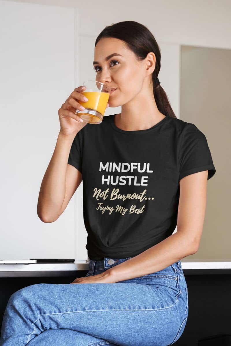 office humor t shirt with a woman wearing a black t-shirt with text 'Mindful Hustle Not Burnout... Trying My Best', holding a glass of orange juice.