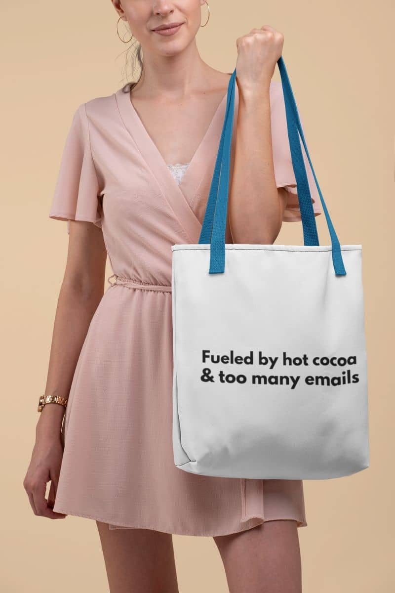 office humor tote bag with a woman holding a tote bag with text 'Fueled by hot cocoa & too many emails' against a beige background
