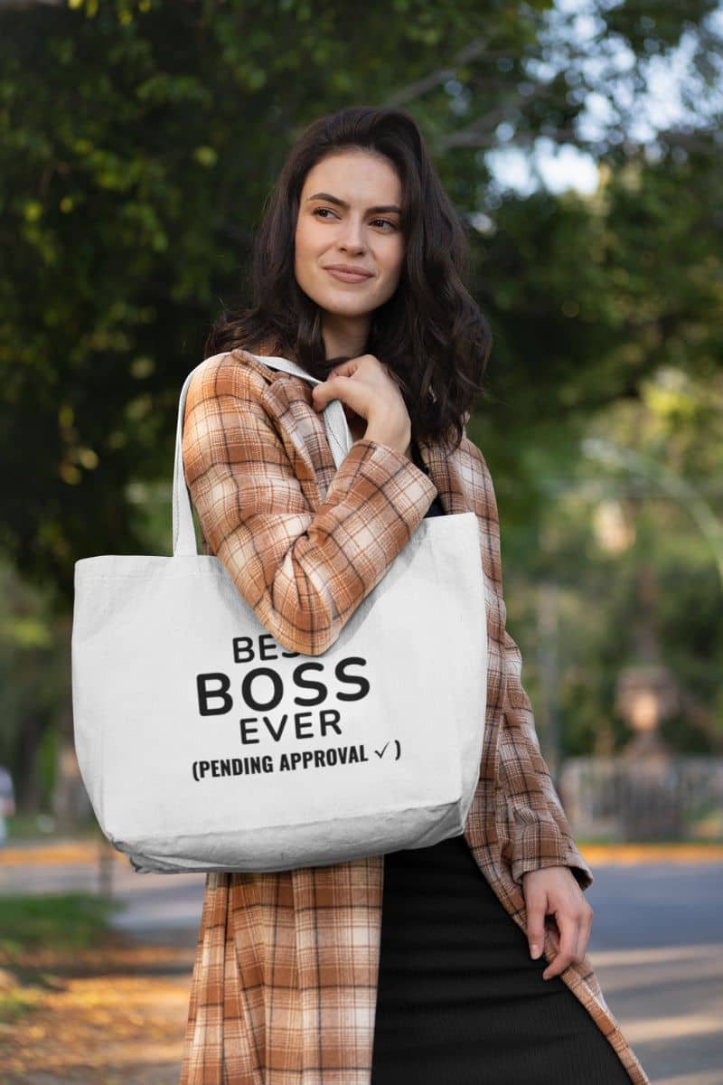 office humor tote bag with a woman holding a tote bag with 'Best Boss Ever (pending approval)' text outdoors.