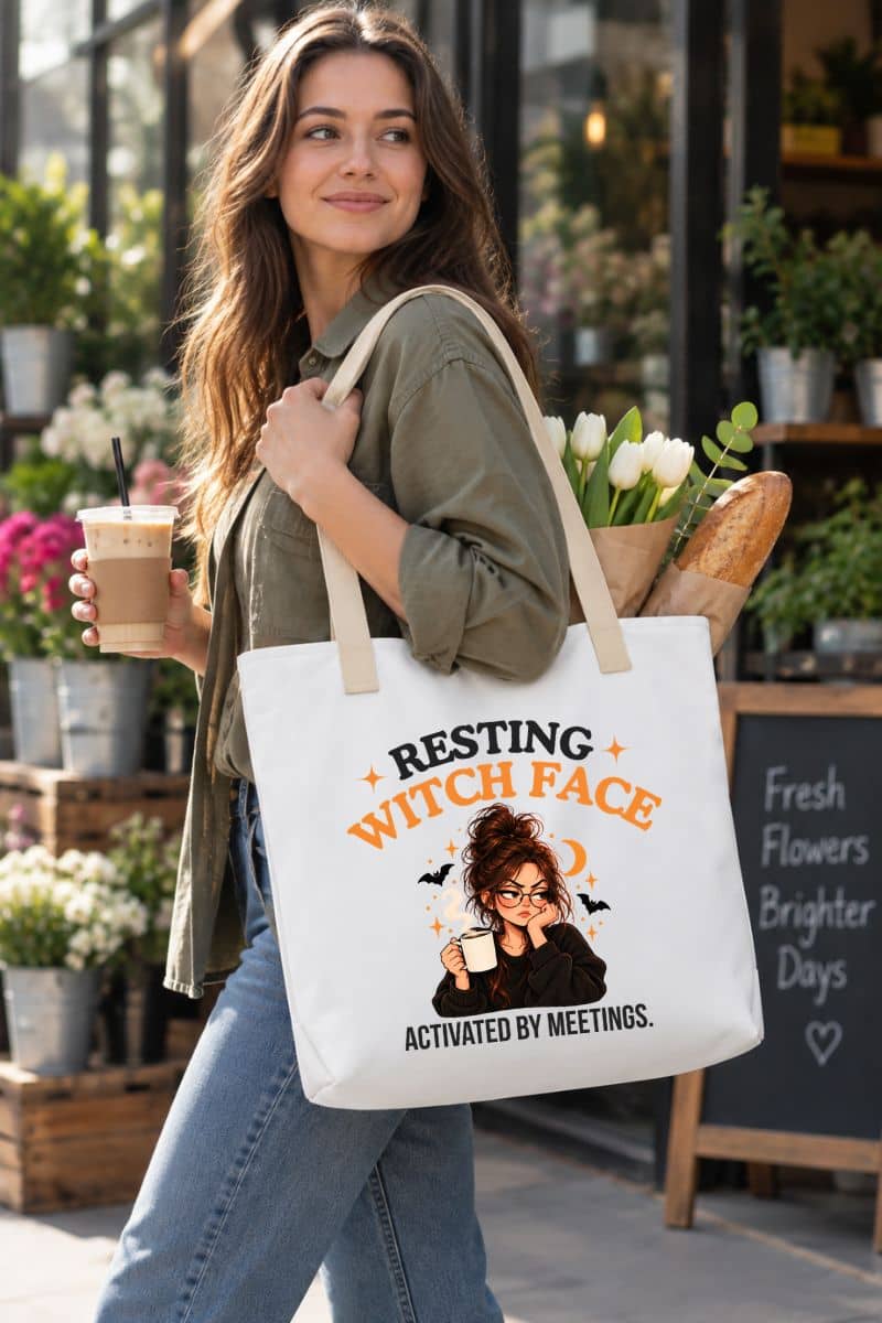 A woman in an olive green shirt and blue jeans carries a white office humor tote with a graphic of a witch and the text 'Resting Witch Face. Activated by Meetings.' in a cafe setting.