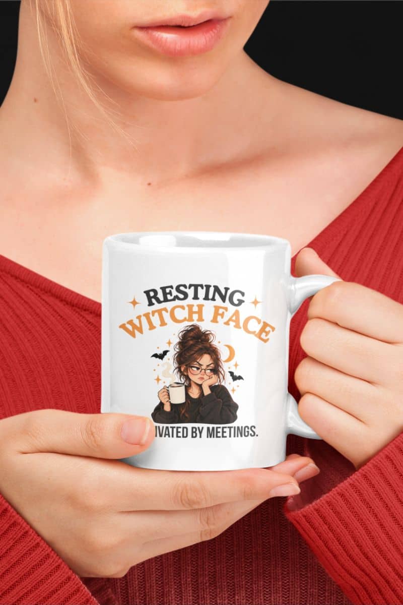A person in a red ribbed sweater holding a office meeting mug with a graphic that reads 'Resting Witch Face. Activated by Meetings.' against a dark background.
