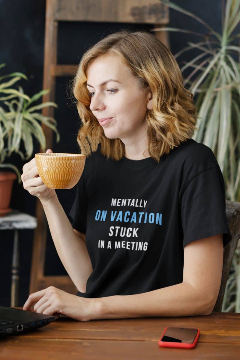 office meeting shirt featuring a woman holding a mug with text on her shirt, sitting at a table with plants in the background