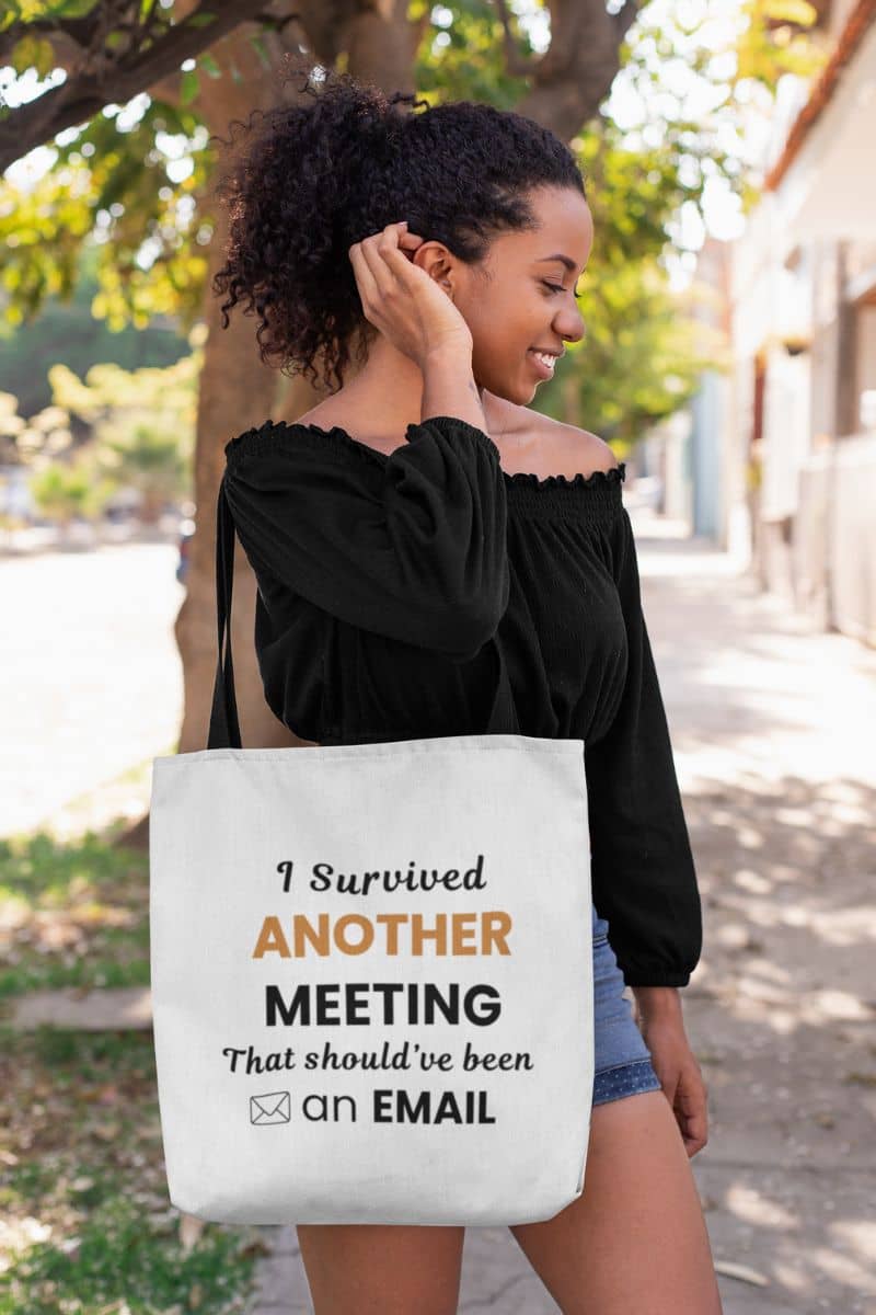 office tote bag with a woman holding a tote bag with text 'Office Humor Tote – I Survived Another Meeting That Should’ve Been an Email' outdoors
