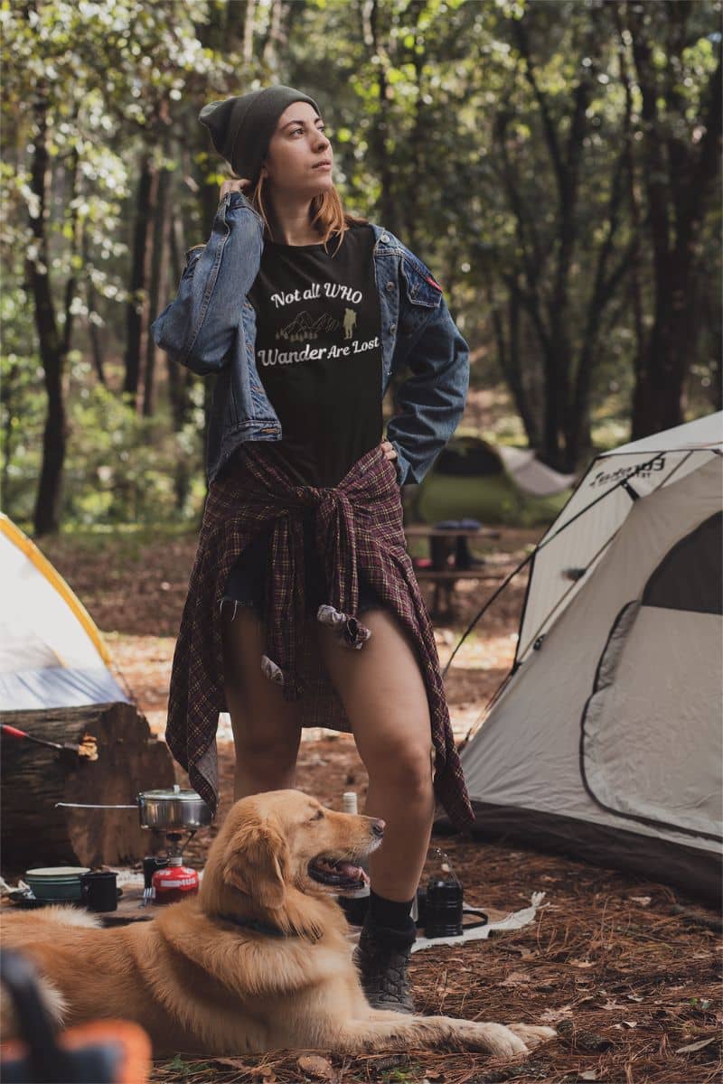 outdoor adventure shirt with a woman wearing at-shirt with text 'Not all who wander are lost', standing with a dog near tents in a forest setting