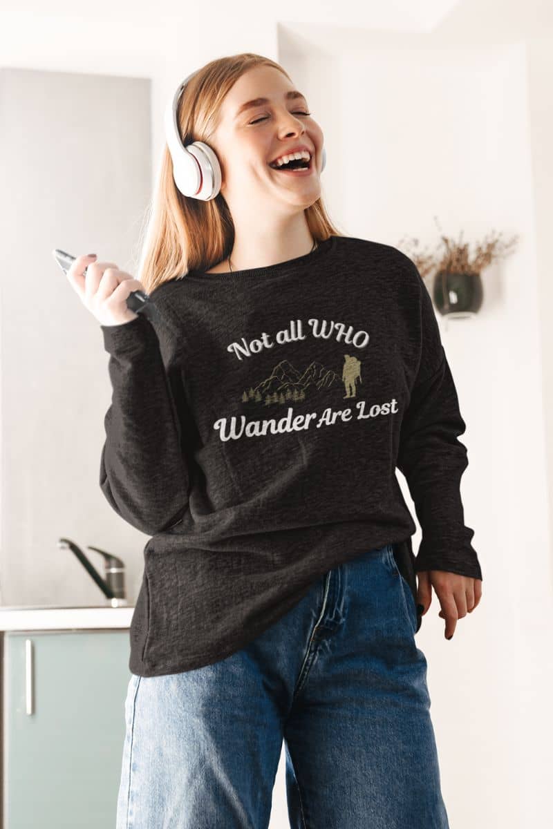 outdoor adventure sweatshirt with a person wearing a sweatshirt with text 'Not all who wander are lost' and headphones, standing in a kitchen.