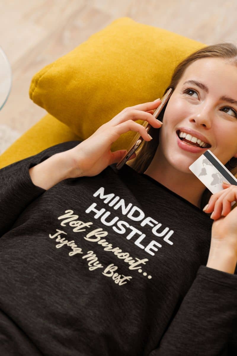 overwhelmed sweatshirt with a woman wearing a black sweatshirt with motivational text 'Mindful Hustle – Not Burnout… Trying My Best', holding a phone and a card.
