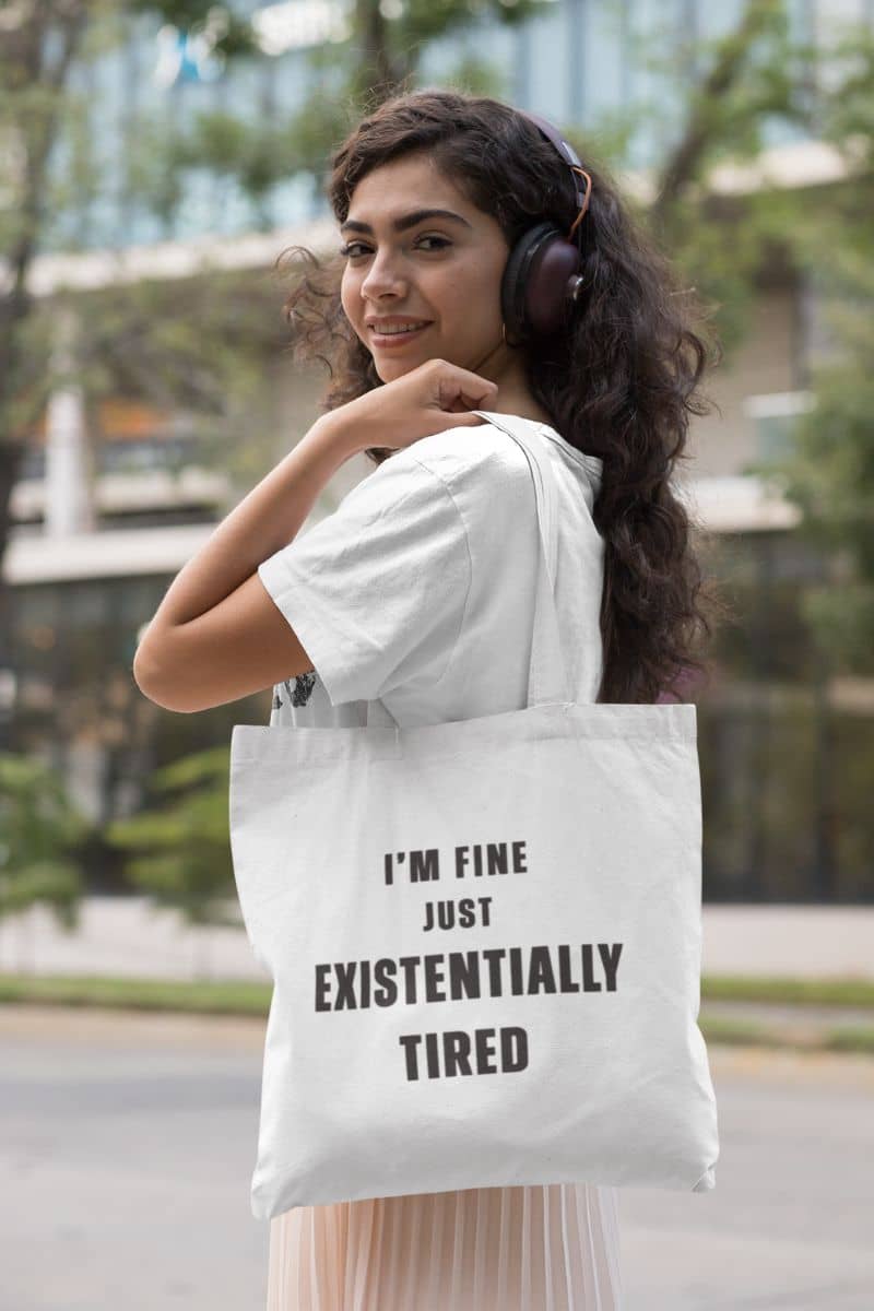 overworked employee tote with a woman holding a tote bag with text 'I’m fine. Just existentially tired.' in an urban setting