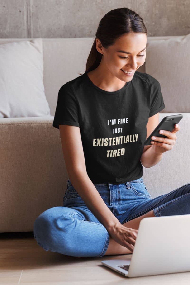 overworked office shirt with a woman wearing a black t-shirt with text 'I’m fine. Just existentially tired.', sitting on the floor with a laptop and phone.