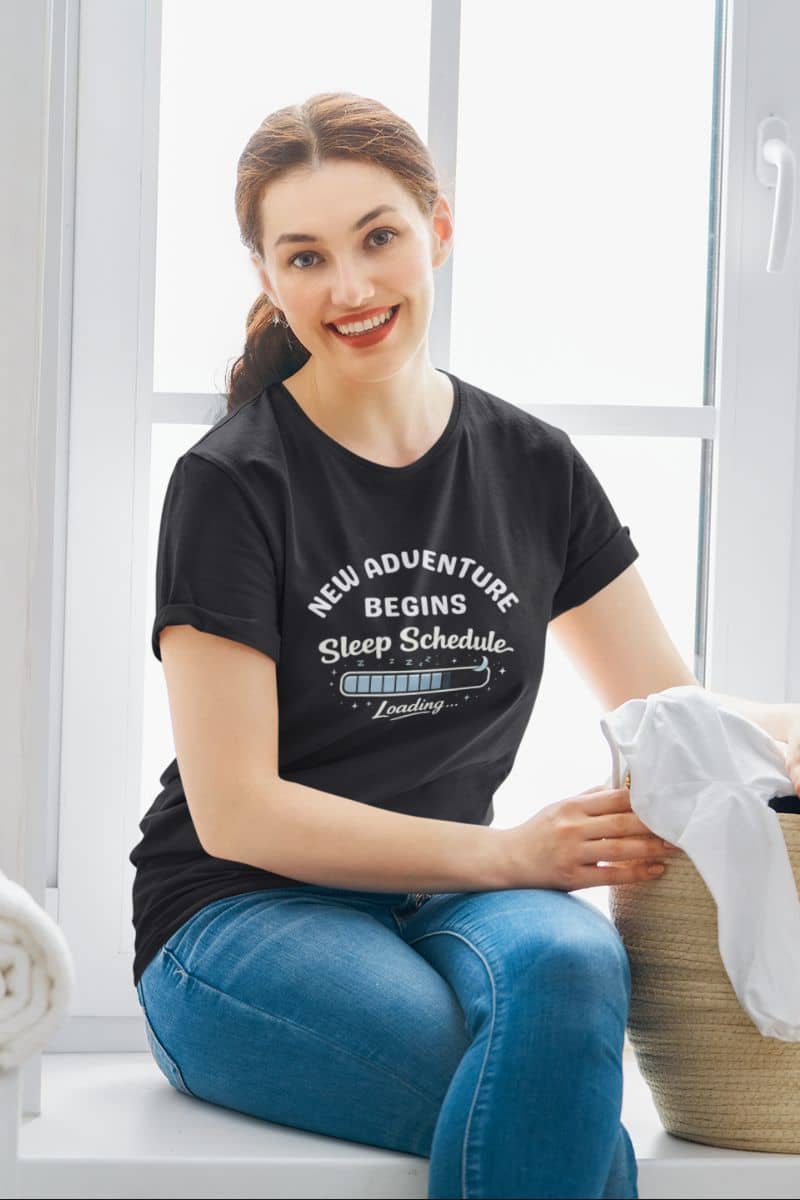 parent life humor shirt with a woman wearing a black t-shirt with text 'New Adventure Begins, Sleep Schedule Loading…', sitting by a window.