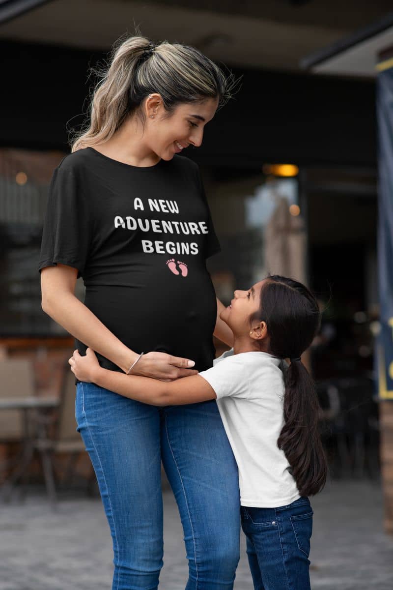 parenthood t-shirt with a pregnant woman wearing a black t-shirt with 'A New Adventure Begins' text, standing with a child.