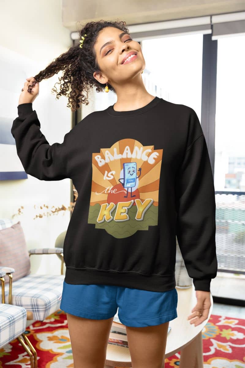 positive affirmation sweatshirt with a woman wearing a black sweatshirt with 'Balance is the Key' graphic design in a living room.