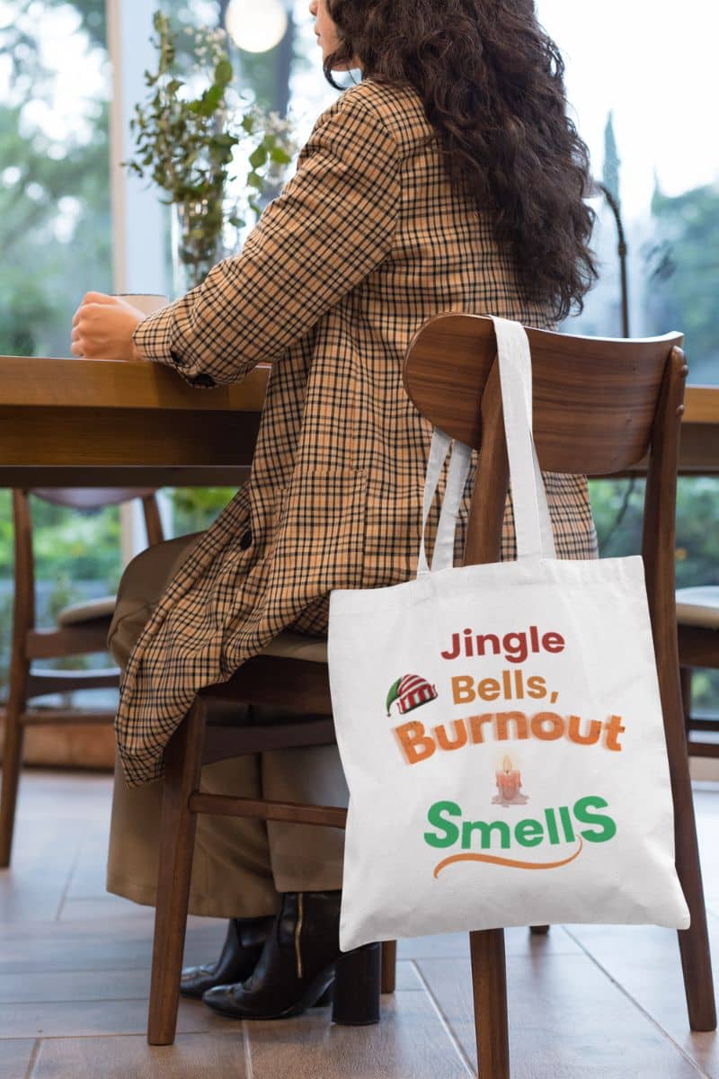 productivity gift tote with a person sitting at a table with a tote bag featuring humorous text 'Jingle Bells, Burnout Smells'.