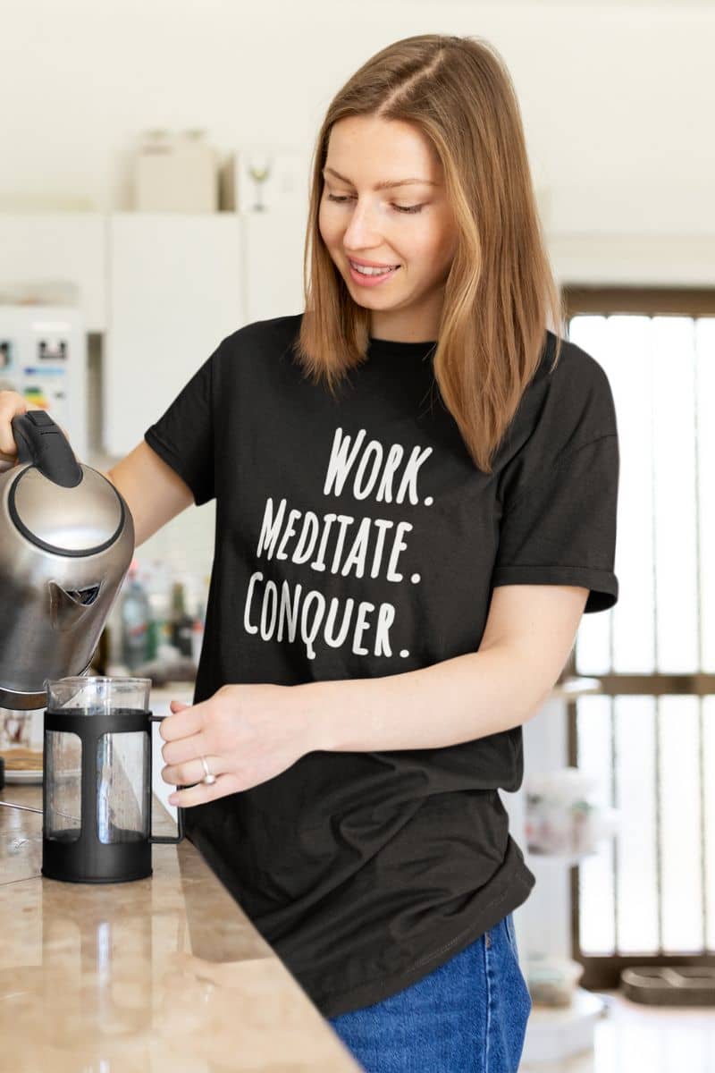 productivity mindset shirt with a woman in a kitchen wearing a black t-shirt with motivational text 'Work. Meditate. Conquer.', holding a kettle.