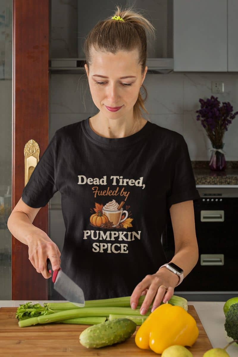 pumpkin shirt with a woman in a kitchen wearing a black graphic t-shirt that reads 'Dead Tired, Fueled by Pumpkin Spice' with an illustration of pumpkins and a mug.