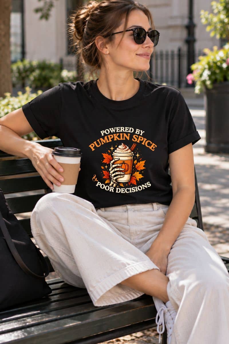 A woman sitting outdoors wearing a pumpkin shirt featuring a "Powered by Pumpkin Spice & Poor Decisions" graphic and a cup of coffee.