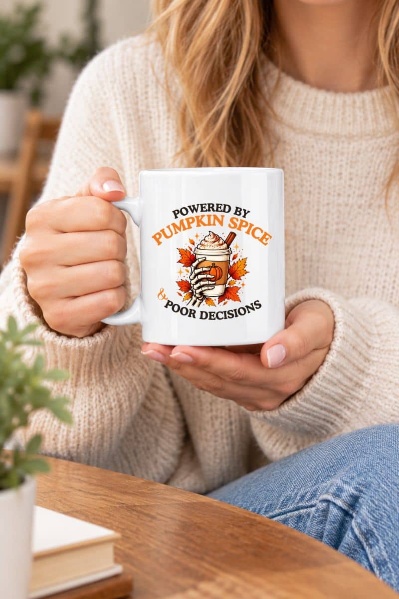 A woman holding a pumpkin spice mug featuring a 'Powered by Pumpkin Spice & Poor Decisions' graphic and a cup of coffee