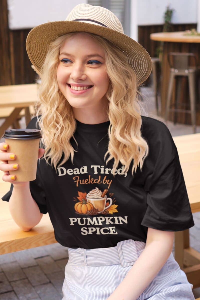 pumpkin spice t shirt with a woman wearing a straw hat and a black graphic t-shirt that reads 'Dead Tired, Fueled by Pumpkin Spice' with a pumpkin and hot cocoa illustration, holding a coffee cup in an outdoor cafe setting.