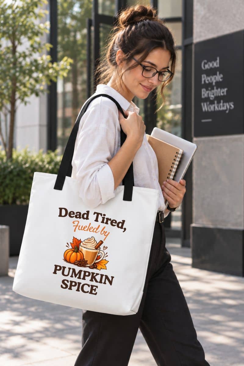 pumpkin spice tote bag with a woman walking outdoors carrying a white canvas tote bag with black handles and a graphic that reads 'Dead Tired, Fueled by Pumpkin Spice' with a pumpkin and spice illustration.