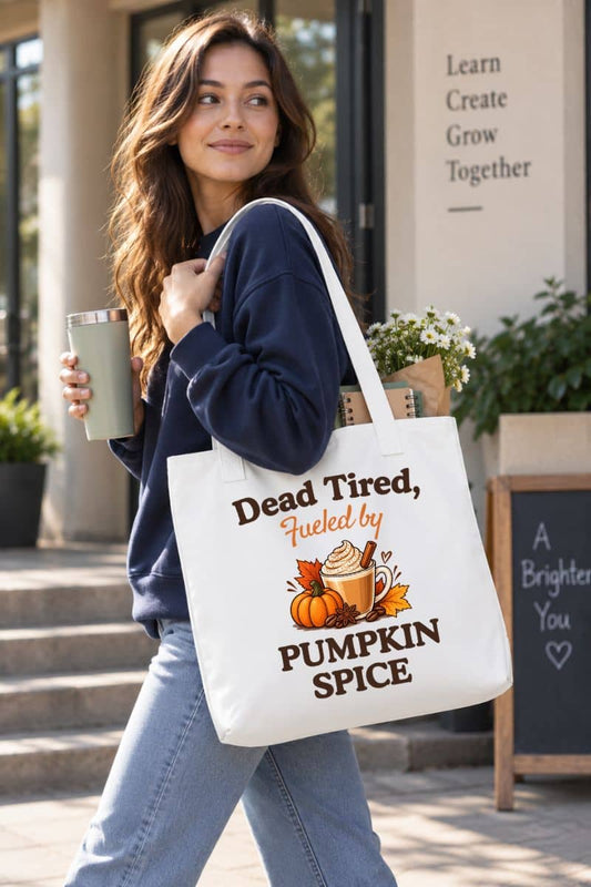 pumpkin tote bag with a woman walking outdoors carrying a white tote bag that reads 'Dead Tired, Fueled by Pumpkin Spice' and a small pumpkin illustration. 
