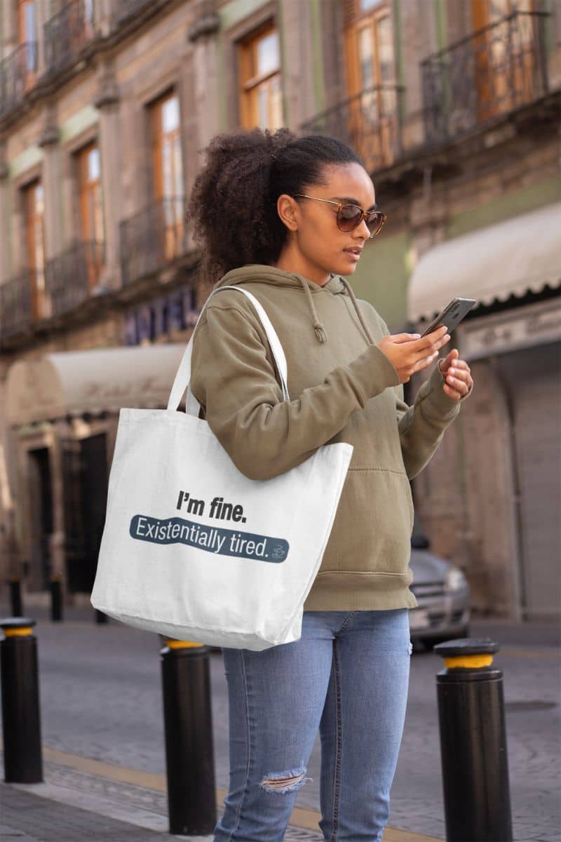 quirky shopper bag with a woman holding a tote bag with humorous text 'I’m fine. Just existentially tired' in an urban setting