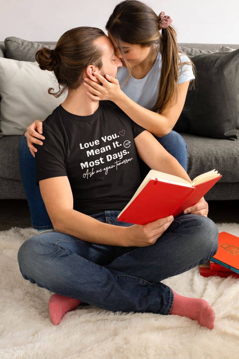 real love humor shirt with a man wearing a black t-shirt with a message 'Love You. Mean It. Most Days. Ask me again tomorrow.', sitting on the floor with a woman, and reading a red book.