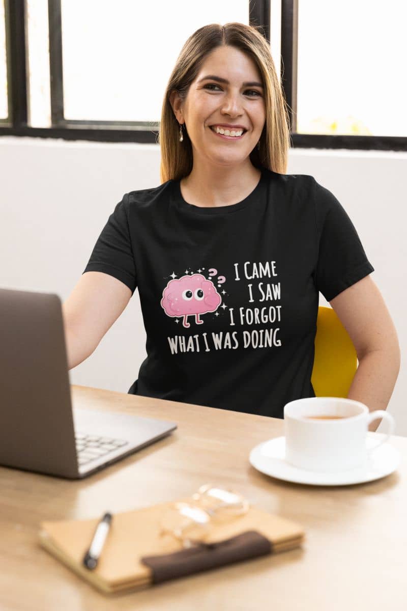 relatable humor t-shirt with a woman wearing a black t-shirt with a graphic and text 'I came, I saw, I forgot what I was doing', sitting at a desk with a laptop and coffee.